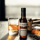 A 5oz bottle of Hella Cocktail Co. Cocktail Bitters: Aromatic (Certified Non-GMO) rests on a wooden table beside two amber drinks, surrounded by spices and warm lighting—an ideal way to elevate your favorite classic cocktails.