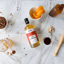 A bottle of Hella Cocktail Co. Old Fashioned Mix rests on white tiles, surrounded by an orange peel bowl, bitters bottle, a cocktail glass, sugar cubes, spices, and a wooden muddler.