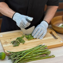 A person wearing NULU Safety Gloves by NULU Inc., which offer Level 5 cut resistance, chops asparagus on a wooden cutting board with a curved knife to demonstrate proper kitchen safety. A lime and a wooden bowl are visible nearby.