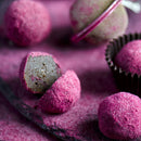 Close-up of truffles rolled in SALTOPIA Chocolate Sugar: HEYO COCOA 16oz, one sliced to show its creamy center. In the background sits a tea infuser dusted with the same SALTOPIA Infused Sea Salts pink chocolate sugar.