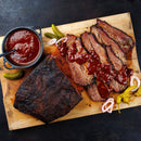 Sliced barbecued brisket topped with SALTOPIA Infused Sea Salts' Saltopia Hickory Salt: CABIN FEVER and barbecue sauce, served on a wooden board with pickles, onions, and extra sauce on the side.
