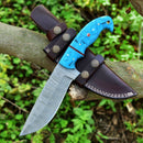 The Texan Knives Custom Handmade Damascus Steel Hunting Knife features a 10-inch patterned blade and resin handle, resting on a tree branch with its leather sheath nearby amid green foliage.