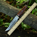 The Texan Knives Damascus Steel Hunting Knife features a 10" patterned blade, brass guard, resin pakkawood handle, and comes with a leather sheath. Shown on a tree branch next to a ruler amid green foliage.