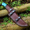The Texan Knives Custom Handmade Damascus Skinning Knife – 4.5" Blade features a blue handle and comes sheathed in brown leather with silver rivets, displayed on logs amidst green leaves and plants.