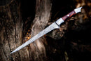 The Handmade Damascus Steel Fish Fillet Knife by Texan Knives, featuring a 13” patterned blade, rosewood handle, and leather sheath, is displayed outdoors resting against a weathered tree trunk.