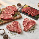 A selection of premium raw steaks from the Dude Food High Steaks Box by Dude Food, featuring T-bone, Kansas City Strip Steaks, and ribeye cuts, displayed on a marble surface with herbs, salt, pepper, and lemon slices—all set for preparation.