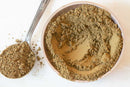 A bowl filled with World Spice Hojicha Powder, a light brown roasted green tea powder from Japan by World Spice, sits on a white surface with a metal spoon holding a scoop of the same powder placed beside it.