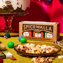 A box of the Spicewalla 3 Pack Holiday Baking Collection by Spicewalla, featuring festive blends such as Apple Pie Spice and Pumpkin Spice, sits on a table with a plate of mixed nuts in front. Colorful felt balls, a matchbox, and part of a dessert appear in the vibrant and festive background.