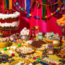 A festive table featuring the Spicewalla 3 Pack Holiday Baking Collection by Spicewalla, surrounded by desserts, nuts, candies, ribbon and pompom garlands, dominoes, dice, pops of colorful spices like Apple Pie Spice and Pumpkin Spice from the collection, all set against a pink backdrop to create a vibrant celebration scene.