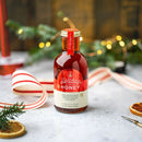 A bottle of Honey Truck Co Holiday Honey® from Honey Truck Co rests on a table adorned with festive decorations—ribbon, evergreen branches, dried orange slices, and twinkling lights in the background—making it the perfect infused honey Christmas gift.