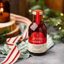 A glass bottle of Honey Truck Co. Holiday Honey® – Christmas Spiced Honey with a red and cream label, featuring the text Holiday Infused Honey and a description of cinnamon, clove, vanilla, and nutmeg flavors.
