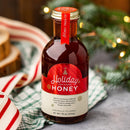 A glass bottle labeled Honey Truck Co Holiday Honey® rests on a cloth, surrounded by festive ribbon and softly blurred pine branches with twinkle lights in the background, making it the perfect Christmas gift for anyone who loves infused honey from Honey Truck Co.