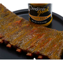 A rack of ribs coated in Gourmet Carolina Gold Honey Carolina Gold BBQ Sauce, positioned next to a bottle of the same mustard-based sauce on a black platter.
