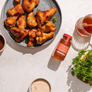 A plate of honey-glazed chicken wings sits beside a jar of Honey Truck Co Raw Datil Pepper Infused Honey, a glass of raw honey with a dipper, a bowl of powder, and a bunch of fresh parsley on a light-colored table.