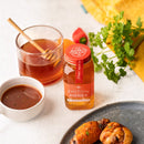 A bottle of Honey Truck Co Raw Datil Pepper Infused Honey stands by a glass of tea with a honey dipper, a bowl of sauce, fresh herbs, and a plate of cooked chicken. Crafted by Honey Truck Co with raw honey and spicy Datil peppers, this distinctive blend delivers sweet heat to every dish.