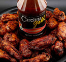 A bottle of Gourmet Carolina Gold Hot Vinegar Pepper Carolina Gold BBQ Sauce surrounded by a pile of glazed chicken wings.