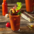 A Bloody Mary cocktail garnished with celery, shrimp, olives, cherry tomatoes, pickles, pepper, and herbs is served in a glass rimmed with SALTOPIA Hot Honey infused sea salt, set on a rustic wooden table.