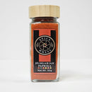 A glass jar with a wooden lid, branded Spice Wheel Hungarian Paprika, contains 50g of vivid red sweet paprika powder. The label showcases the signature circular Spice Wheel logo and a row of five colorful dots, reflecting the lively flavors characteristic of Hungarian cuisine.