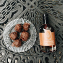 A glass plate with five chocolate-covered treats sits beside a bottle of Under The Bark Syrup Co Bourbon + Cinnamon Maple Syrup, its orange label adding elegance to the ornate black metal table.
