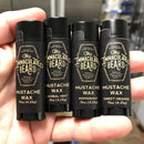 Four tubes of The Immaculate Beard Mustache Wax in herbal mint, peppermint, and sweet orange varieties are held in a person’s hand. Made with natural ingredients, each tube features a black label with gold text and is ideal for shaping a classic handlebar mustache.