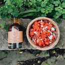 A bottle of Under The Bark Syrup Co Habanero Maple Syrup sits on a stone surface beside a wooden bowl of diced watermelon topped with crumbled white cheese, surrounded by green plants and leaves.