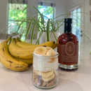 A jar with granola, sliced bananas, and yogurt sits on a countertop beside fresh bananas and a bottle of Under The Bark Syrup Co Bourbon + Cinnamon Maple Syrup. A leafy plant and bright windows are visible in the background.