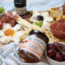A charcuterie board features assorted cheeses, meats, crackers, grapes, and a jar of Terrapin Ridge Farms Hot Pepper Peach Jam in the foreground. A striped cloth is partially visible at the bottom.