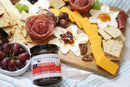 A charcuterie board with crackers, cheddar cheese, salami roses, grapes, pecans, and ricotta with jam is displayed. In front sits a jar of Terrapin Ridge Farms Hot Pepper Bacon Jam by Terrapin Ridge Farms on a striped cloth.
