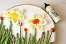A plate with hard-boiled eggs arranged like flowers, cherry tomato flowers, and asparagus stems is served with Terrapin Ridge Farms Dill Pickle Aioli Squeeze—a perfect burger condiment or seafood sauce—beside the plate.