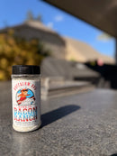 A clear plastic shaker of BuffalowBBQ Bacon Ranch BBQ Rub featuring a label with an illustration of a buffalo wearing a chef’s hat and sunglasses surfing on a wave.