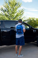 A person wearing a blue Blue Coolers 72 Hour Emergency Kit Essential For 1 Person backpack stands next to a black truck.