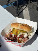 A gluten-free pulled pork sandwich seasoned with BuffalowBBQ Sassy Swine BBQ Rub, served with coleslaw, pickles, and barbecue sauce in a paper food tray.