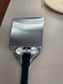 Here’s a rewritten sentence using the provided product data: A close-up of the WPPO Ash Shovel with Black Alumina Handle by WPPO LLC Direct rests on a light-colored surface, its large, flat, shiny metal blade facing upward in the style of a traditional pizza oven ash shovel, with part of a round plate visible in the background.