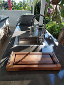 A modern outdoor kitchen showcases a sleek gray countertop with a compact Baby BBQ Board® 18" x 8" cutting board from BBQ Boards®, plus a stainless steel sink and grill. Green plants and trees create an inviting, sunny cooking space.