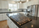 Modern kitchen with white cabinets, stainless steel appliances, black countertops, and a BBQ Boards® Biggest BBQ Board® 36 x 24 Reversible Cutting Board on the island. Pendant lights hang above, with sunlight streaming through the window above the sink.