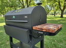 A black pellet grill featuring the BBQ Boards® GMG Peak Prime 2.0 Side Cutting Board stands on green grass in a park, surrounded by leafy trees—ideal for displaying your favorite BBQ Boards® accessories.