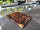 The Beefy BBQ Board® 18" x 15" cutting board by BBQ Boards® with metal handles sits on a shiny outdoor counter near a stainless grill, surrounded by greenery for a welcoming grilling and entertaining space.