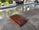 The Basic BBQ Board® 16" x 9" Cutting Board by BBQ Boards® rests on a sleek outdoor kitchen countertop, with a gas grill and sink nearby, and vibrant greenery stretching beyond the cooking area.
