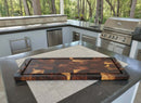 The Burly BBQ Board® 35 x 12 Cutting Board by BBQ Boards® rests on an outdoor kitchen counter amidst stainless steel appliances, gray cabinets, and a built-in grill, with trees visible in the background.