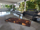 A polished outdoor kitchen features stainless steel appliances, a grill, and the Burly BBQ Board® 35 x 12 Cutting Board from BBQ Boards® on the countertop. Lush greenery and flowering plants can be seen in the background.
