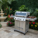 A BBQ Boards® Monument Grills Mesa 405BZ Side Cutting Board is featured on a stainless steel gas grill with four knobs, set on a tiled patio among lush plants, vibrant flowers, and a stone fountain in the background.