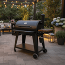 A BBQ Boards® Pit Boss 1150DX Side Cutting Board sits beside a black grill on a stone patio under string lights, surrounded by potted flowers, lanterns, and cushioned seating in a landscaped backyard.