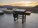 At sunset, a modern patio showcases a black gas grill on wheels with the BBQ Boards® 30" Blackstone Griddle Side Cutting Board, cushioned seating, and a rectangular fire pit. Mountains and a valley complete the scenic backdrop.