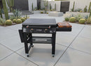 A black flat-top outdoor griddle with a BBQ Boards® 30" Blackstone Griddle Side Cutting Board stands on wheels on a spacious tiled patio, surrounded by cacti, succulents, and a fire pit in modern landscaping.