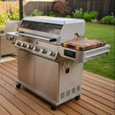 A BBQ Boards® Monument Grills Denali 610 Side Cutting Board is attached to a six-knob outdoor gas grill, which stands on a wooden deck in a backyard with green grass and lush plants.