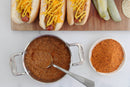 Three hot dogs smothered in chili meat sauce made with World Spice Coney Island Hot Dog Blend and topped with mustard rest on a cutting board next to crisp pickle spears. Below is a pot of rich chili, a spoon, and a bowl of World Spice Coney Island Hot Dog Blend seasoning on a white surface.