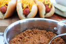 A metal pot filled with hearty chili meat sauce sits in the foreground, while Coney Island hot dogs seasoned with World Spice Coney Island Hot Dog Blend from World Spice, topped with shredded cheddar and a pickle spear, are blurred in the background on a wooden board.