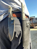 A person in dark pants with a brown belt wears two Messermeister Leather Belt Sheaths for Overland Utility Knife at their waist, knife handles visible. Vehicles and a sunny outdoor background can be seen.