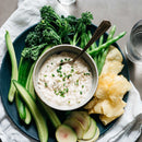 A plate with cucumbers, green beans, broccolini, thinly sliced radishes, potato chips, and a creamy dip topped with chives and SALTOPIA Infused Sea Salts Bacon & Ranch Seasoning: HOWDY DO is served with a glass of water nearby.