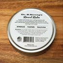 A tin of Dr. B.Strong’s Cutting Board Balm from NULU Inc. rests on a wooden surface. The lid’s label lists its use as a food-safe wood conditioner for protecting cutting boards, along with directions, features, and company details in black text.